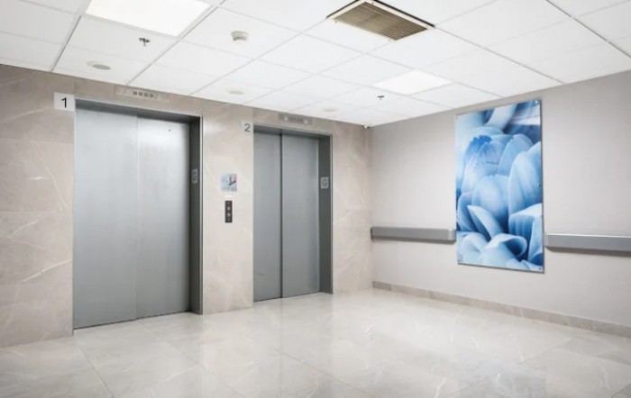 A clean and modern hospital corridor with two elevator doors and a large abstract painting on the wall.