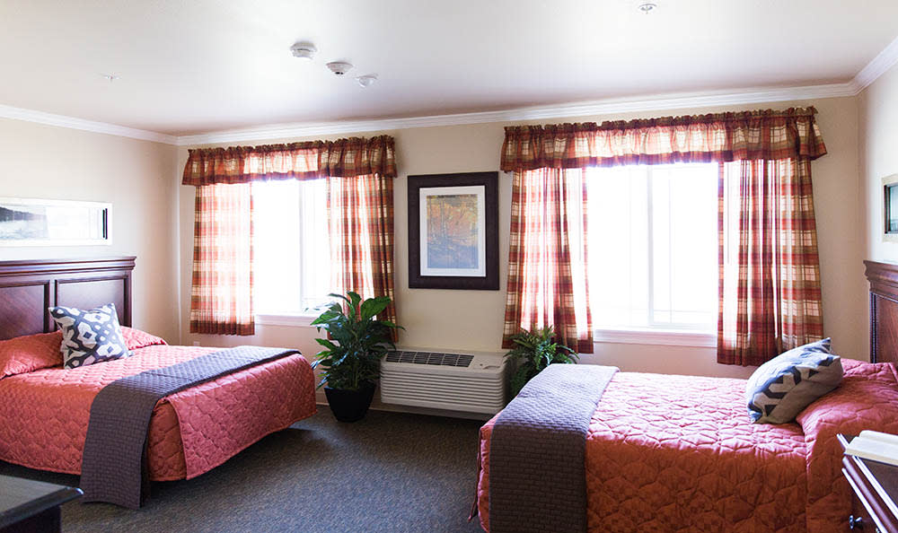 A well-lit hotel room featuring two beds, patterned curtains, a framed artwork, and potted plants.