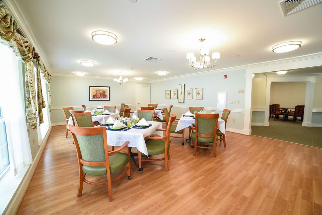 A well-lit dining room with neatly arranged tables and chairs, featuring chandeliers and wooden flooring.