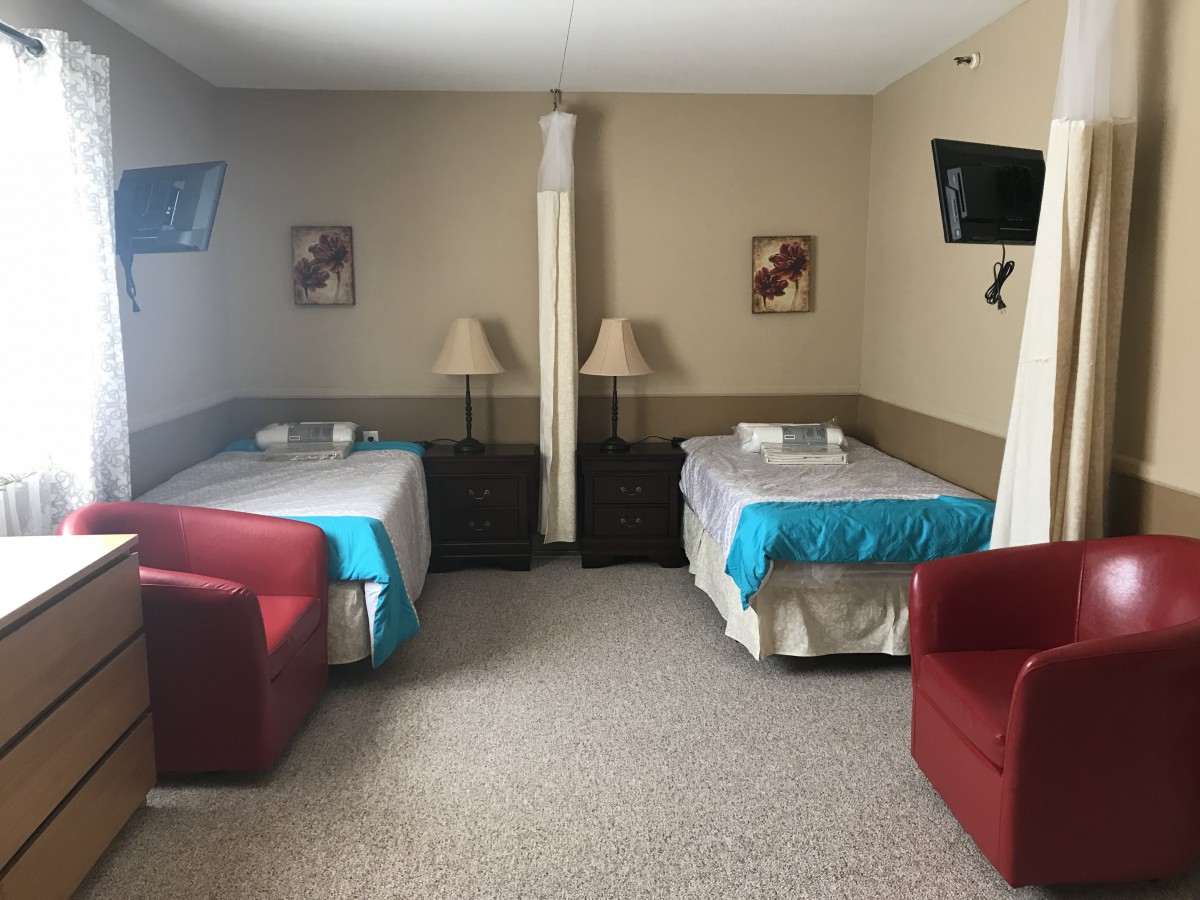 A hotel room with twin beds, featuring cozy seating and wall-mounted televisions.