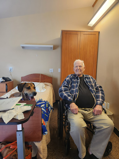 An elderly man in a wheelchair smiles alongside a dog in a care facility room.