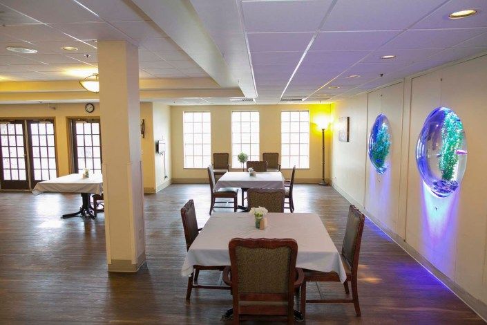 A cozy dining area with neatly arranged tables and chairs, featuring a wooden floor and large windows that allow natural light to brighten the space.