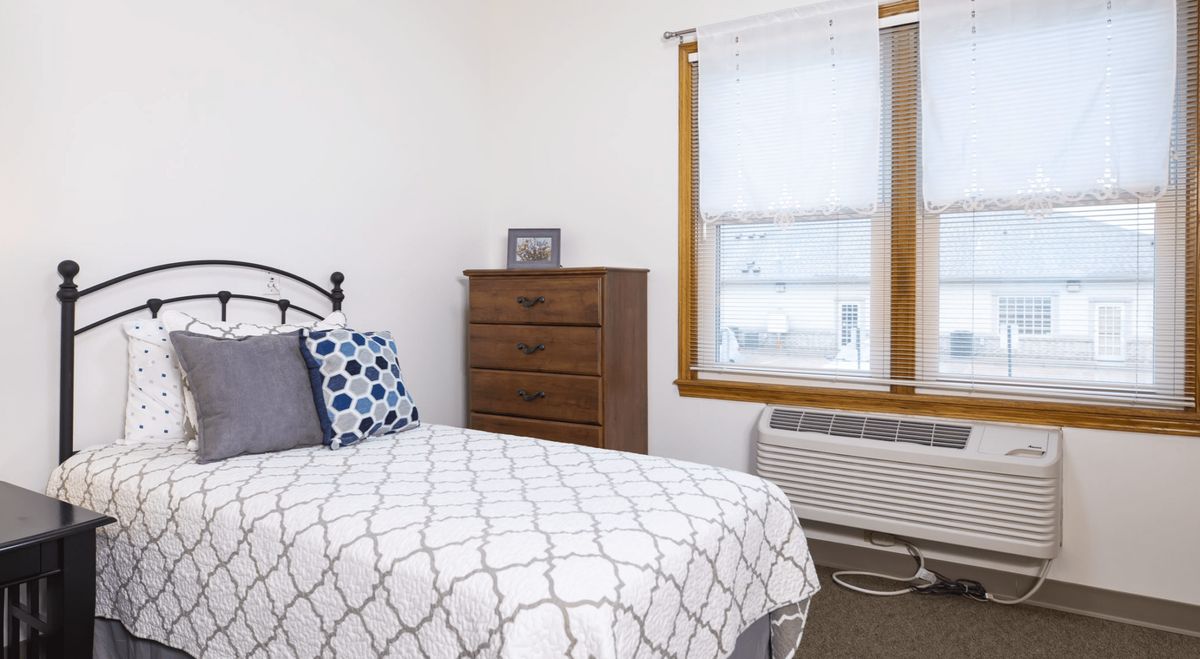 A cozy bedroom with a neatly made bed, a dresser, and a window with an air conditioning unit.