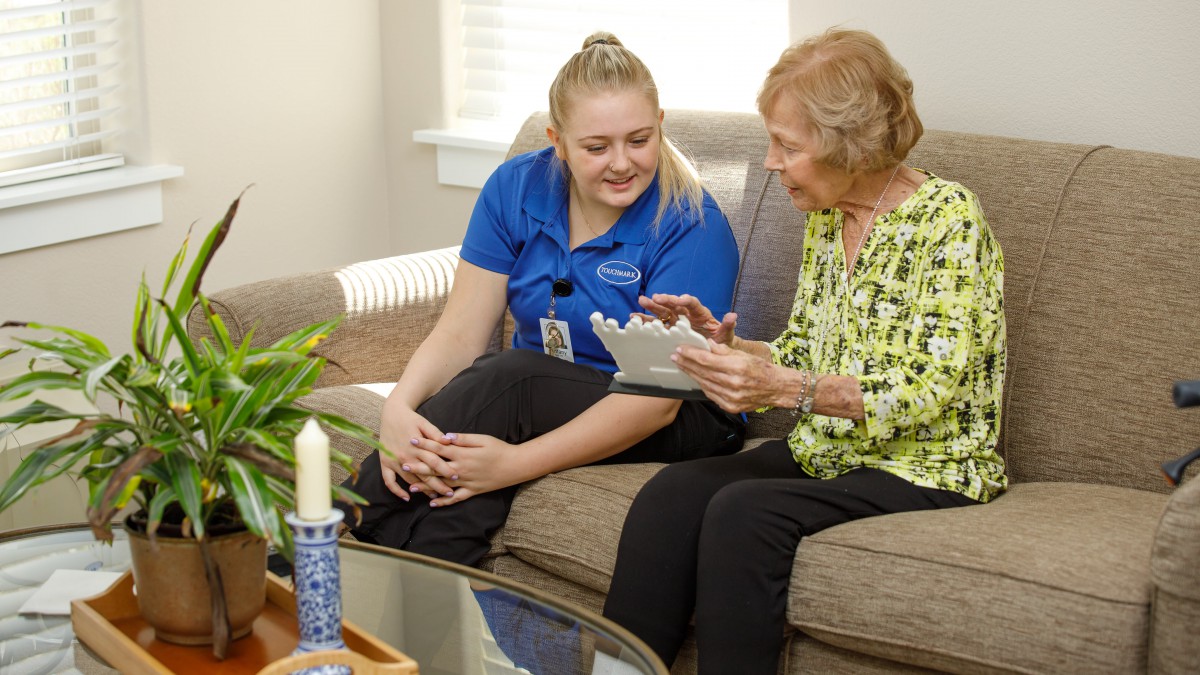 A caregiver sits with an elderly woman, discussing something on a tablet in a cozy living room setting.