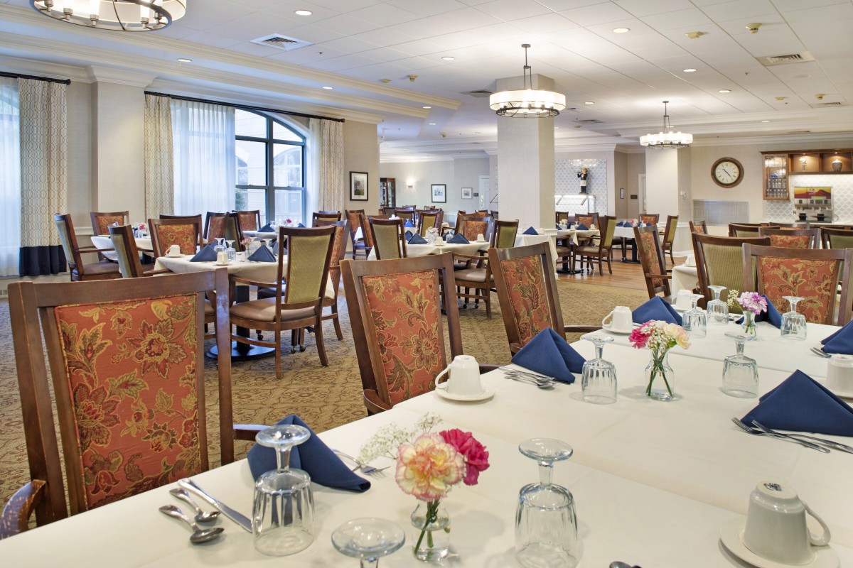 A formal dining room with elegantly set tables and chairs, featuring flowers and napkins.