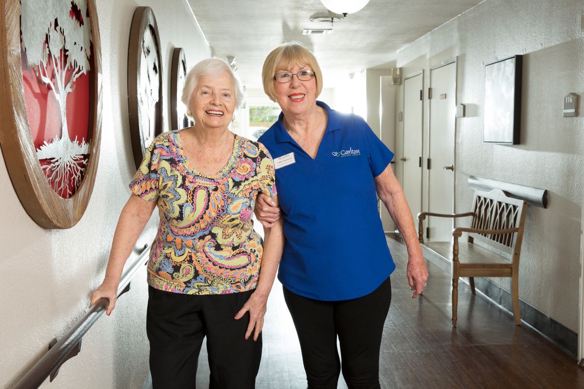 An elderly woman walking with assistance from a caregiver in a nursing home hallway.