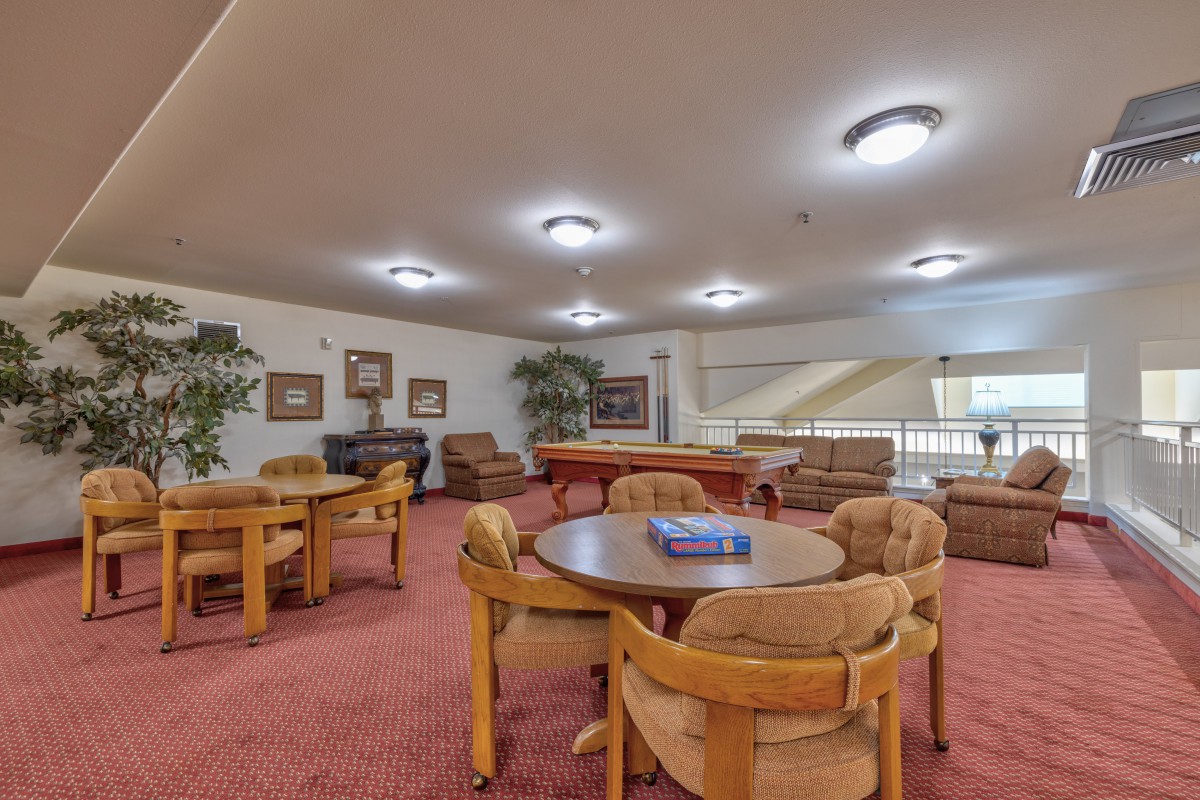 A cozy lounge area with multiple tables and chairs, featuring a carpeted floor and warm lighting.