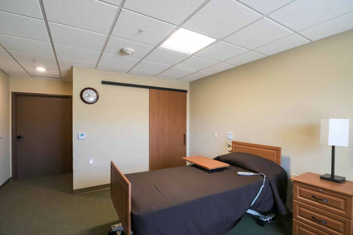 A hospital room with a single bed and an overbed table, featuring a clock on the wall and a wooden sliding door.