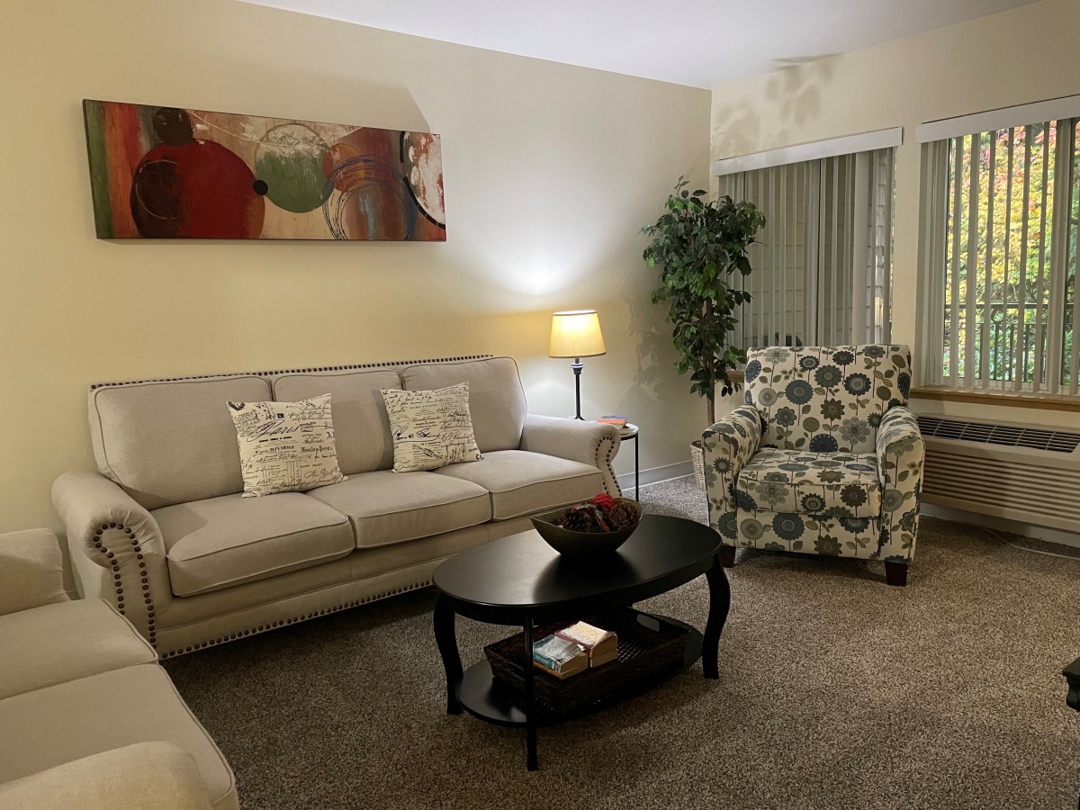 A cozy living room with a sofa, armchair, and coffee table, decorated with artwork and a potted plant.