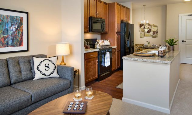 A cozy apartment interior featuring a living room with a sofa and a lamp, adjacent to a kitchen with wooden cabinets.