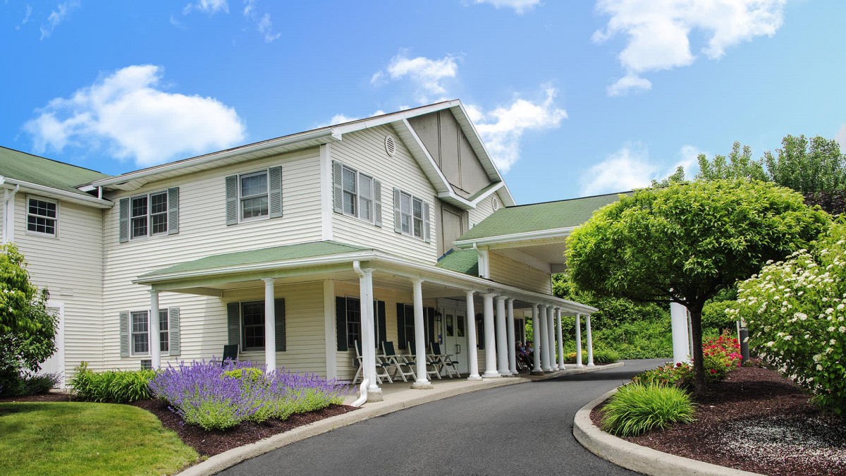 A two-story residential building with a green roof and a covered porch, surrounded by landscaped gardens on a sunny day.