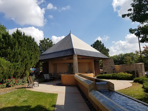 A modern pavilion with a distinctive pyramid roof surrounded by greenery and a flowing water feature.