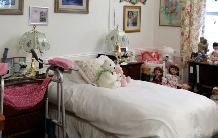 A cozy bedroom featuring a bed adorned with teddy bears and decorative pillows, surrounded by lamps and family photographs.