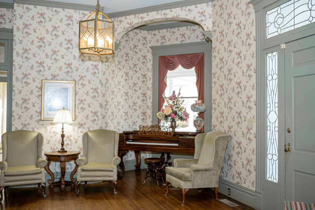 A vintage living room featuring floral wallpaper, a wooden piano, and armchairs.