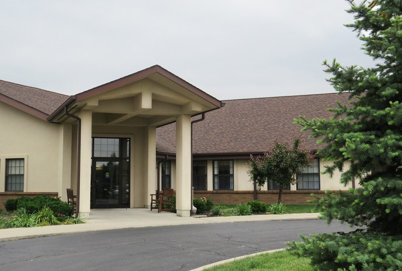 A building with a covered entrance, featuring columns and a well-maintained driveway and surrounding greenery.