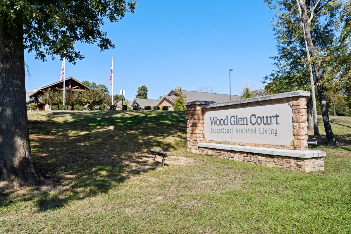 A sign for Hood Glen Court, an assisted living facility, with buildings and a landscaped area in the background.