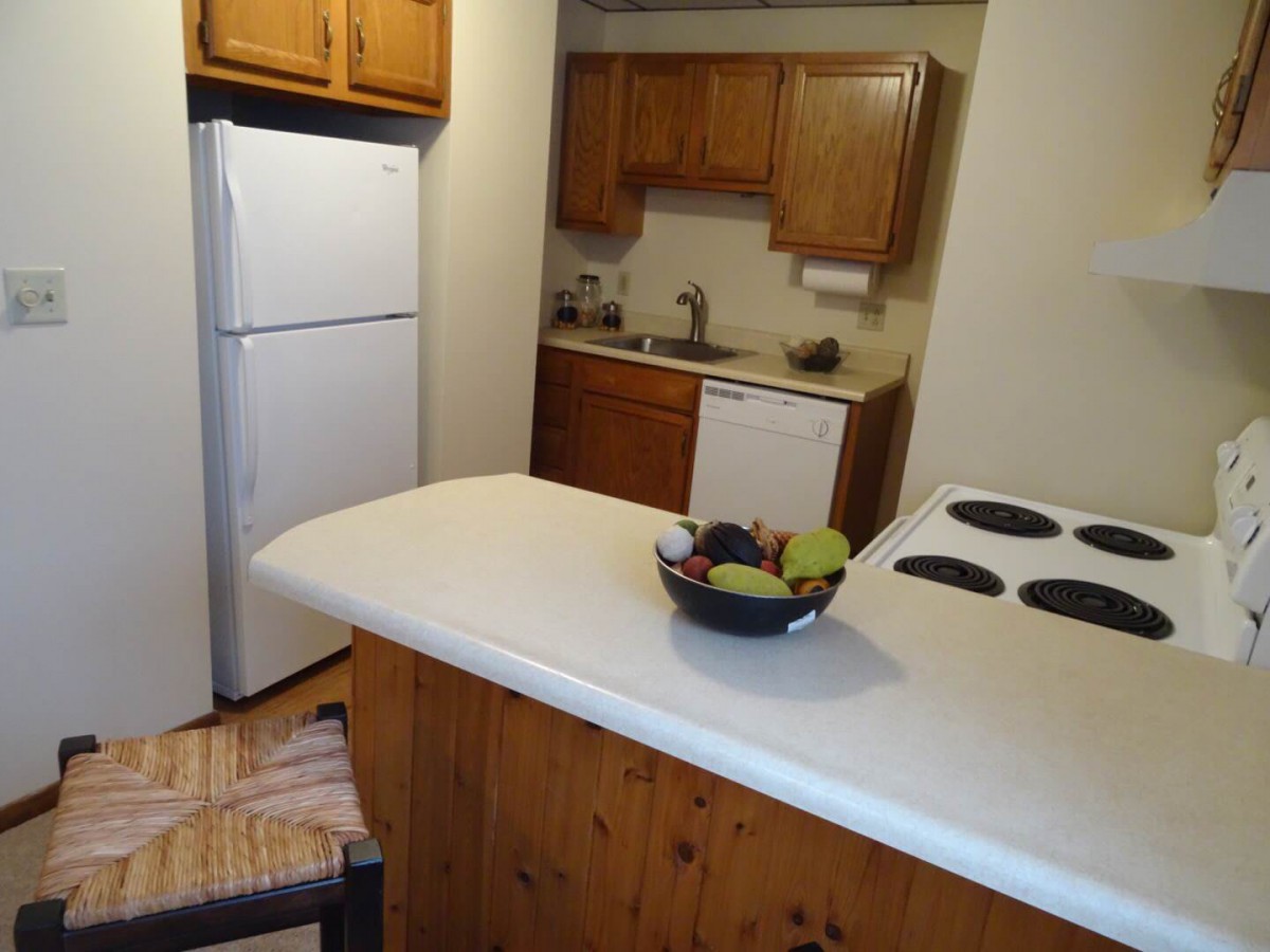 A cozy kitchen with wooden cabinets, a refrigerator, and a countertop with a bowl of fruit.