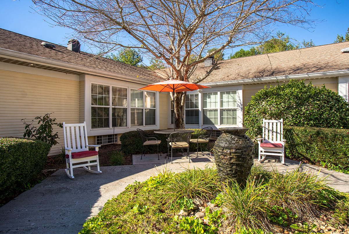 A cozy patio area with rocking chairs and a table under an orange umbrella, surrounded by greenery.