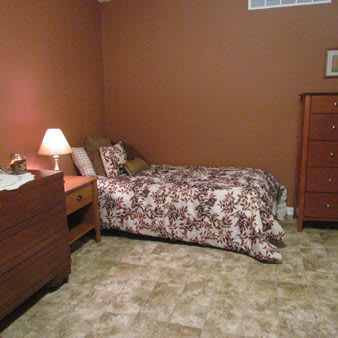 A cozy bedroom with a single bed, nightstand, lamp, and wooden dresser against brown walls.