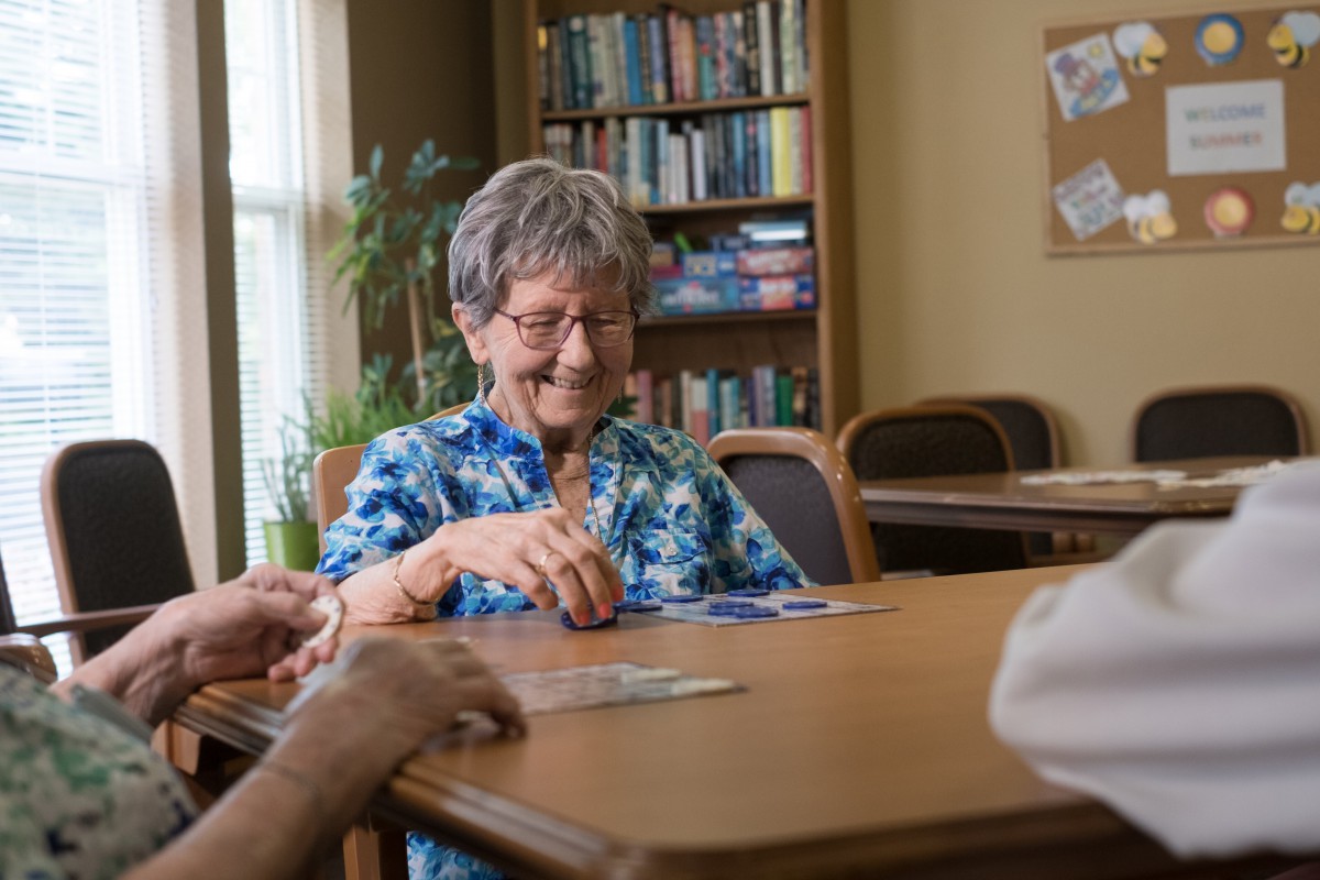 An elderly woman is smiling while playing cards at a table in a cozy room with bookshelves.