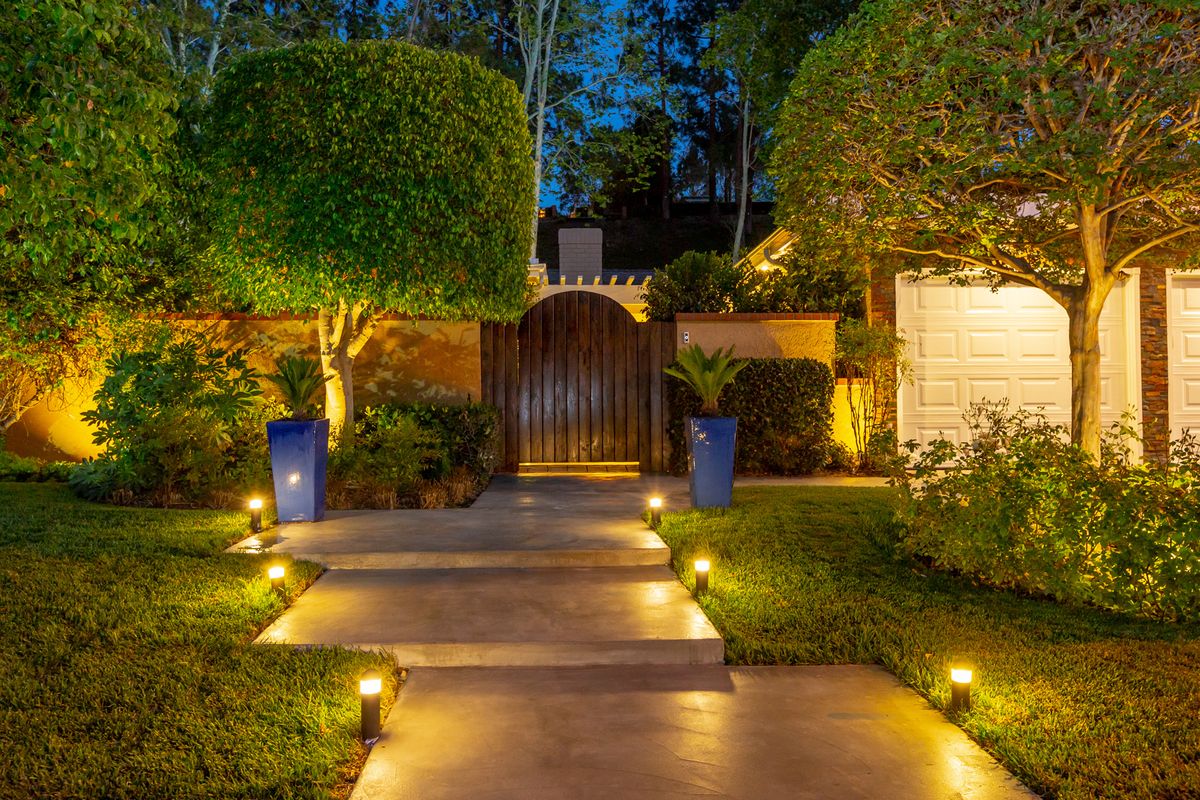 A beautifully lit garden pathway leading to a wooden gate, surrounded by manicured trees and plants at night.