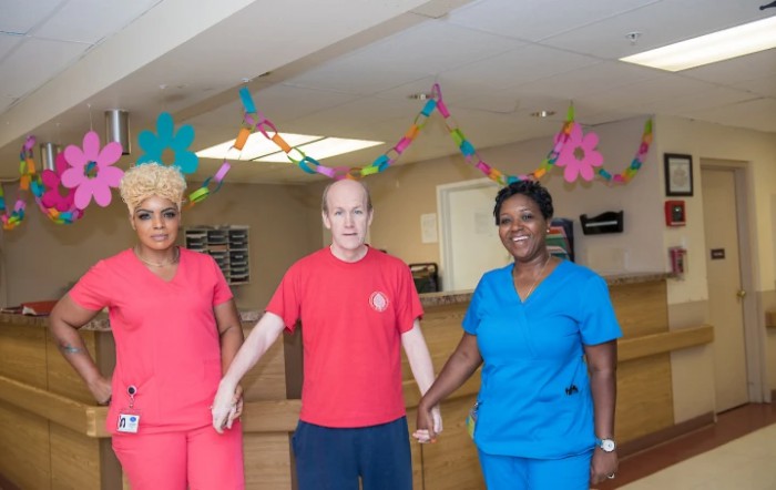 A group of three people holding hands and smiling indoors, with colorful decorations in the background.