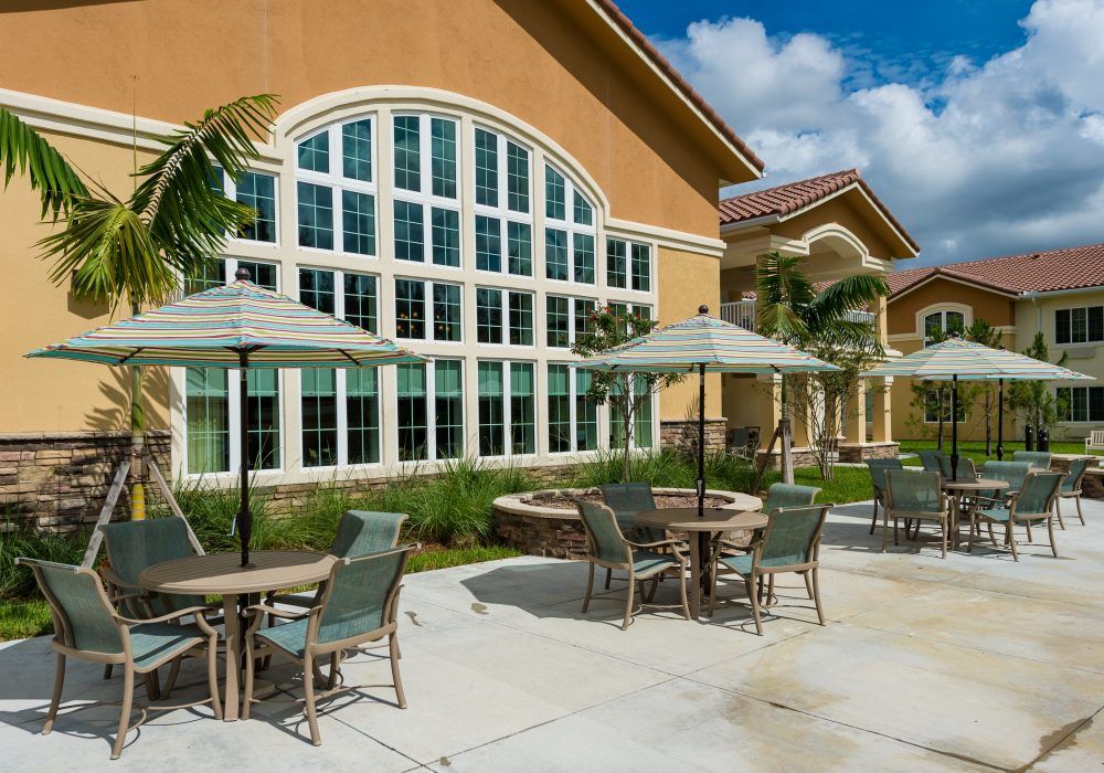 A sunny patio area with tables and umbrellas in front of a building featuring large arched windows.