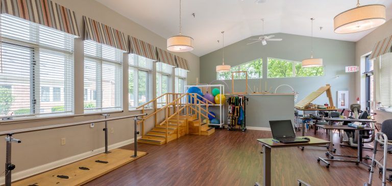 A bright physical therapy room featuring wooden stairs, parallel bars, and various rehabilitation equipment.