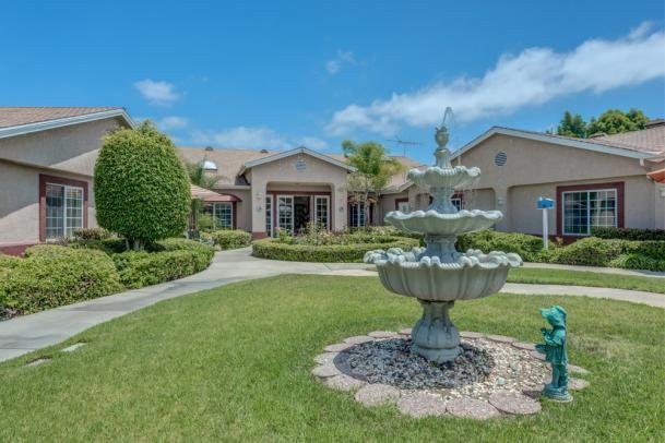 A beautiful garden with a central fountain in front of a large house on a sunny day.