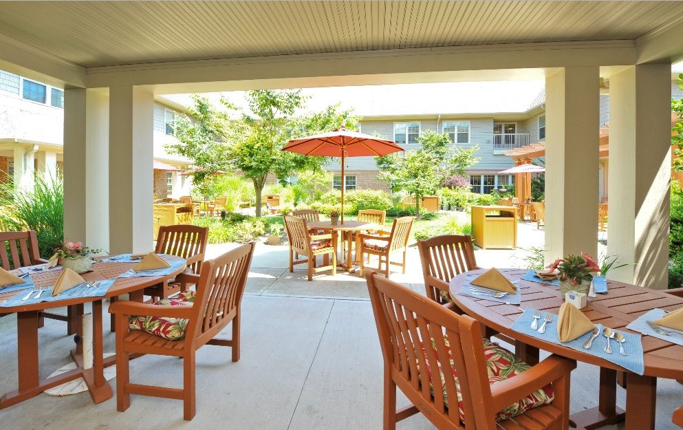 A sunny courtyard with multiple wooden tables and chairs, shaded by umbrellas, surrounded by greenery.