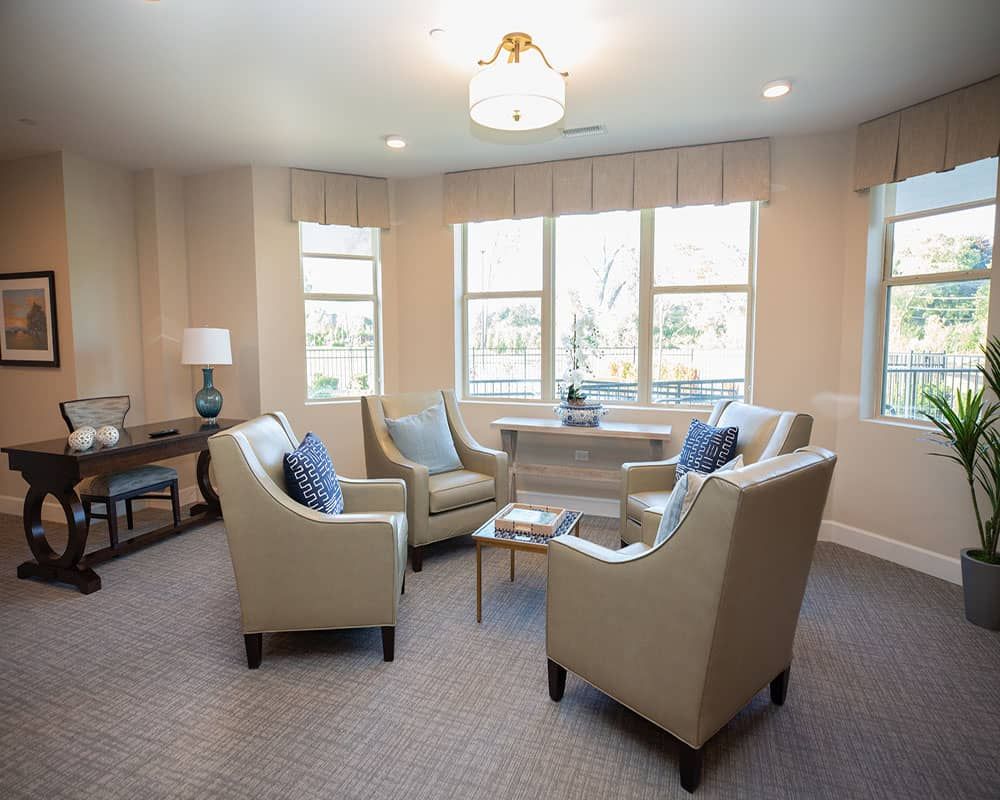 A cozy living room with comfortable chairs arranged around a coffee table, well-lit by natural light from multiple windows.