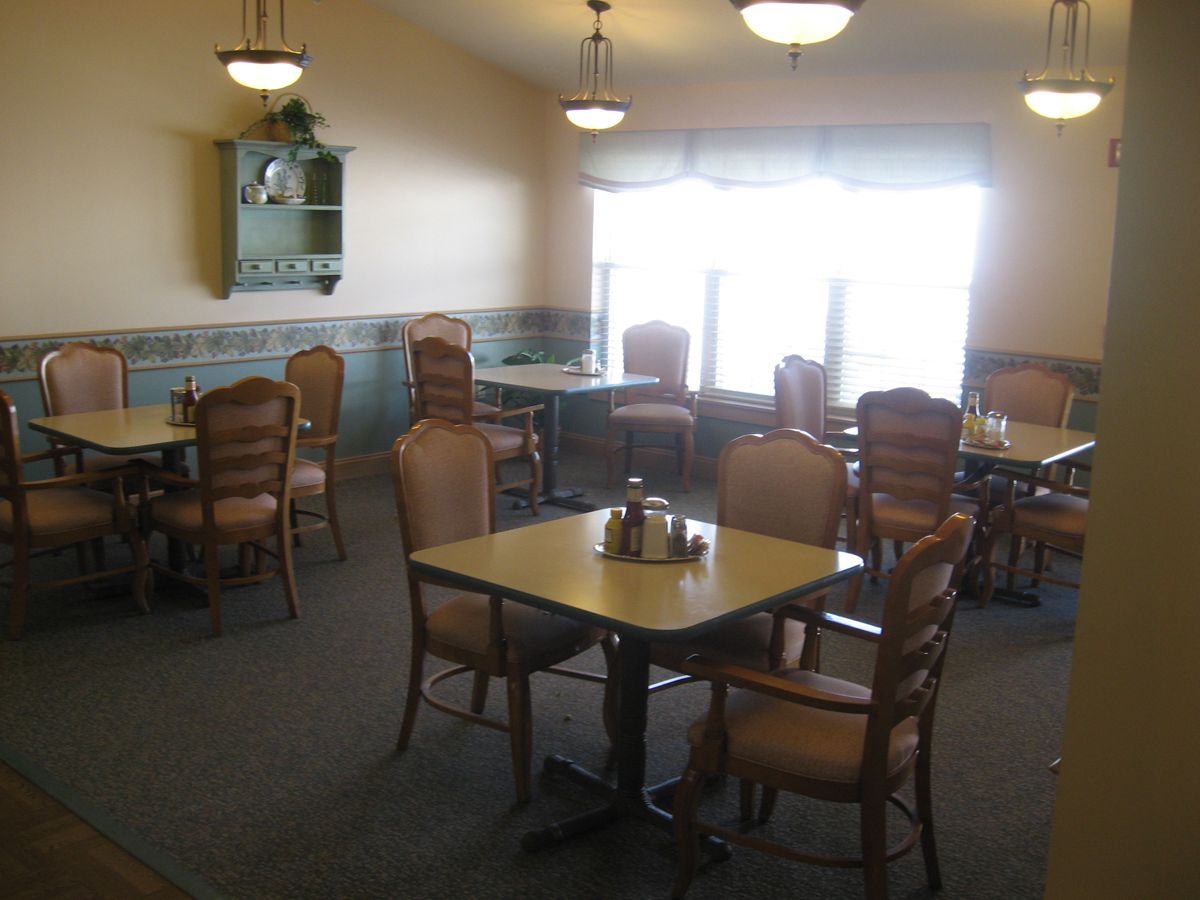 An empty dining room with multiple tables and chairs, featuring large windows and ceiling lighting.