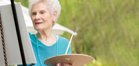 An elderly woman painting outdoors with a palette and brush.