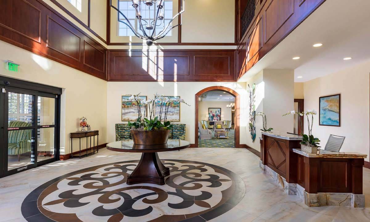 A spacious and elegant lobby featuring a chandelier, wood paneling, decorative flooring, and a central plant display.