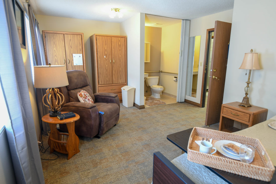 A cozy living room with a comfortable armchair, a wooden table with a lamp, and a wardrobe.