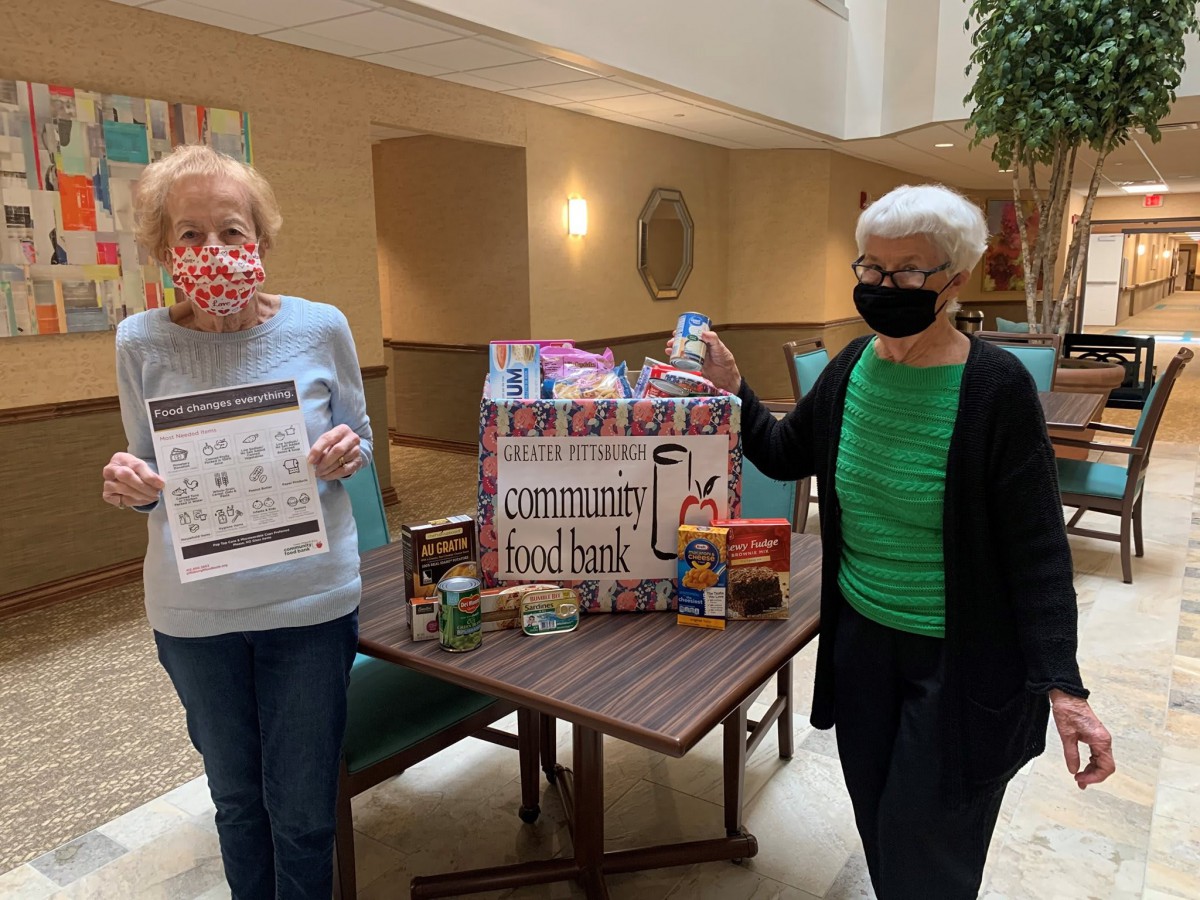Two women stand beside a donation box filled with food items during an indoor event.