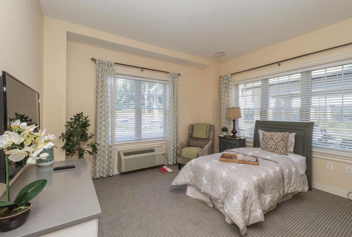 A cozy bedroom with a neatly made bed, two large windows with patterned curtains, a comfortable chair, and modern decor.