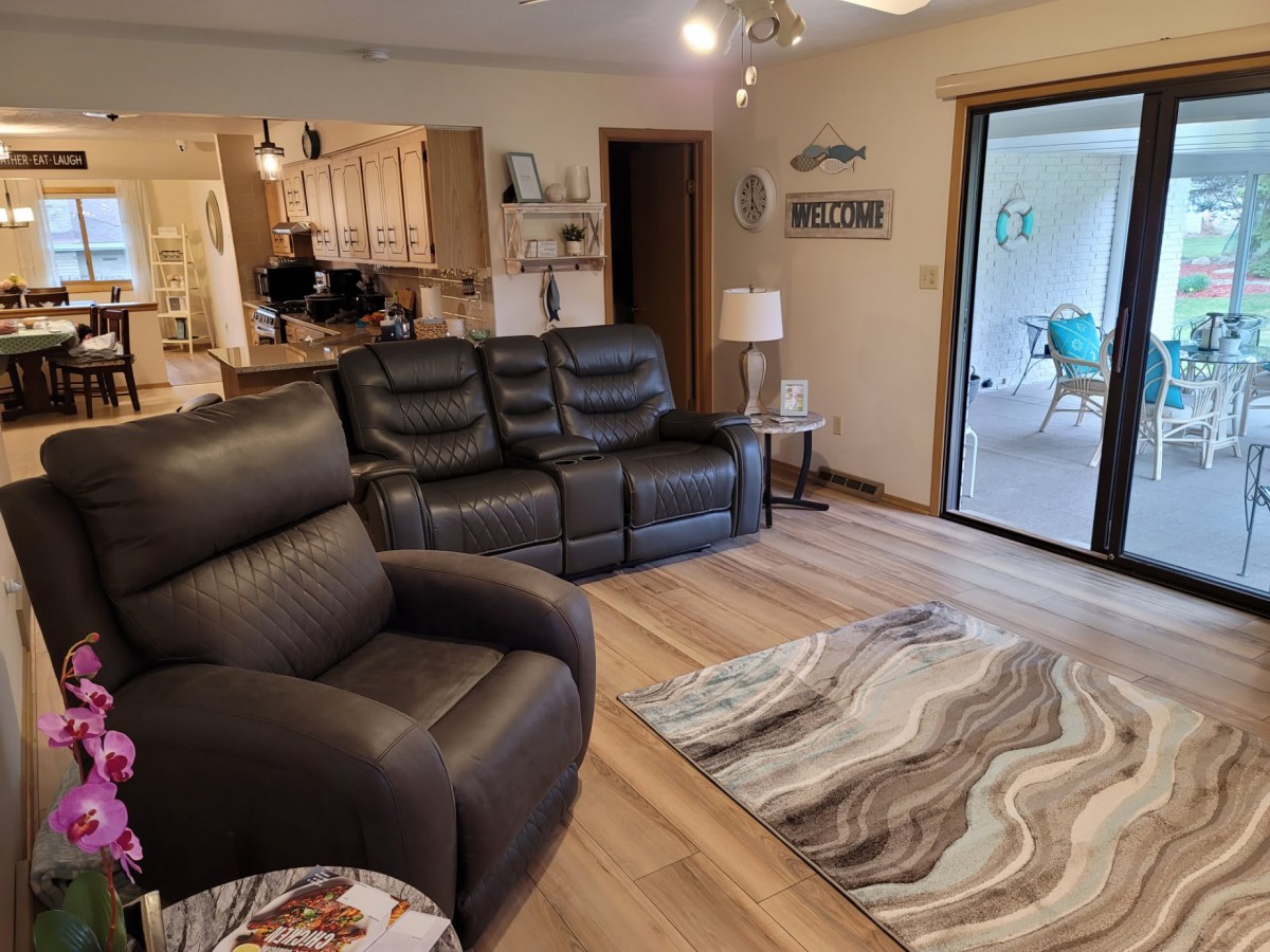 A cozy living room with leather recliners, a patterned rug, and an open view into the kitchen through a wide sliding glass door.