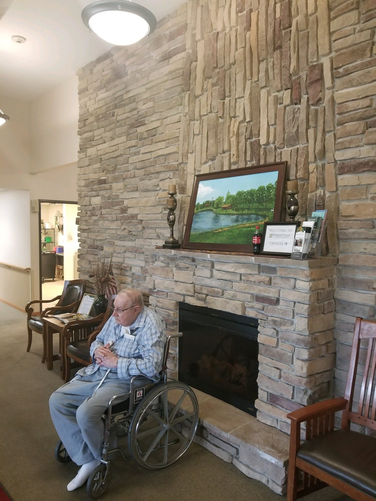 An elderly person in a wheelchair sits near a fireplace adorned with decorations and a painting on the mantel.