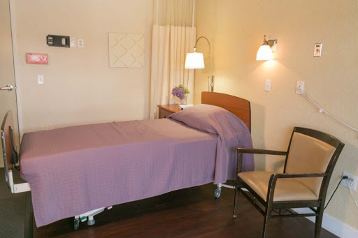 A cozy hospital room featuring a single bed with a purple blanket, a chair, and warm lighting.