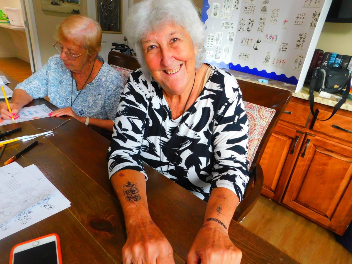 An elderly woman with tattoos smiles while sitting indoors.