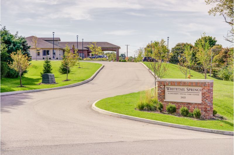 A view of a driveway leading to a building surrounded by greenery, with a sign at the entrance.