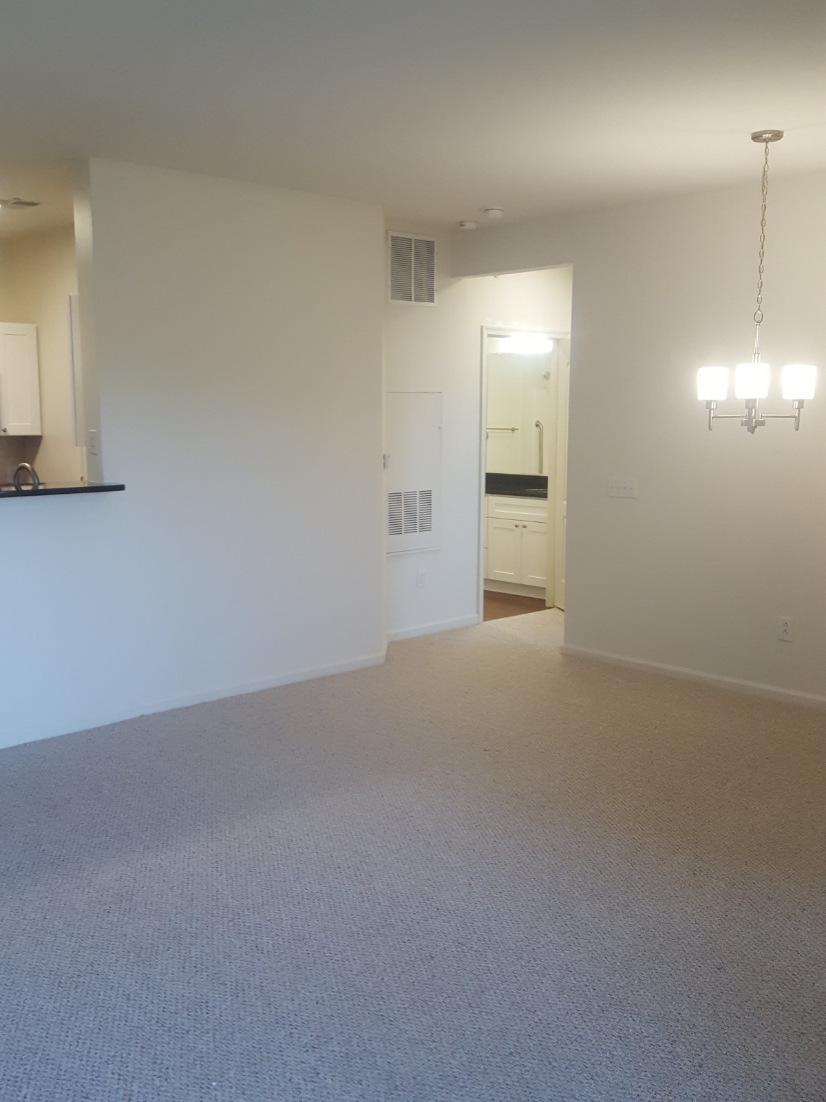 An empty apartment living room with a dining area, featuring a carpeted floor and a chandelier.