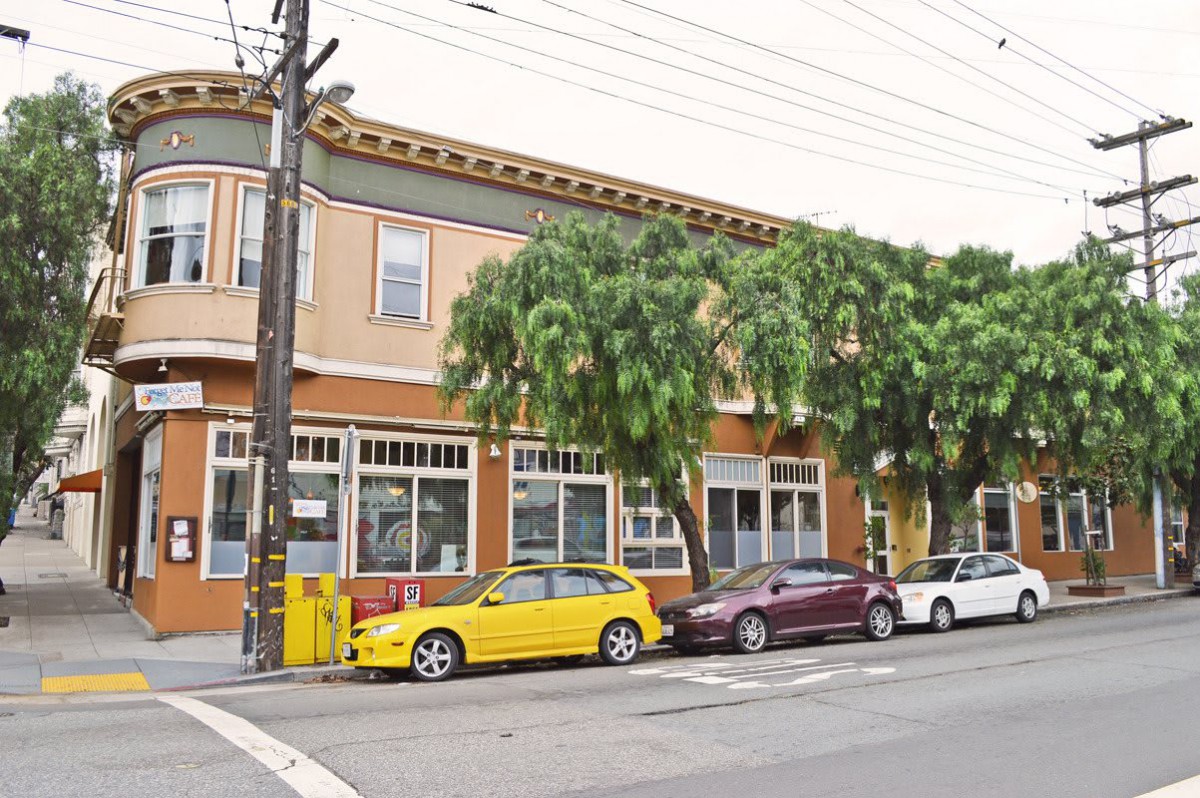 A street view of a two-story building with cars parked in front and trees lining the sidewalk.