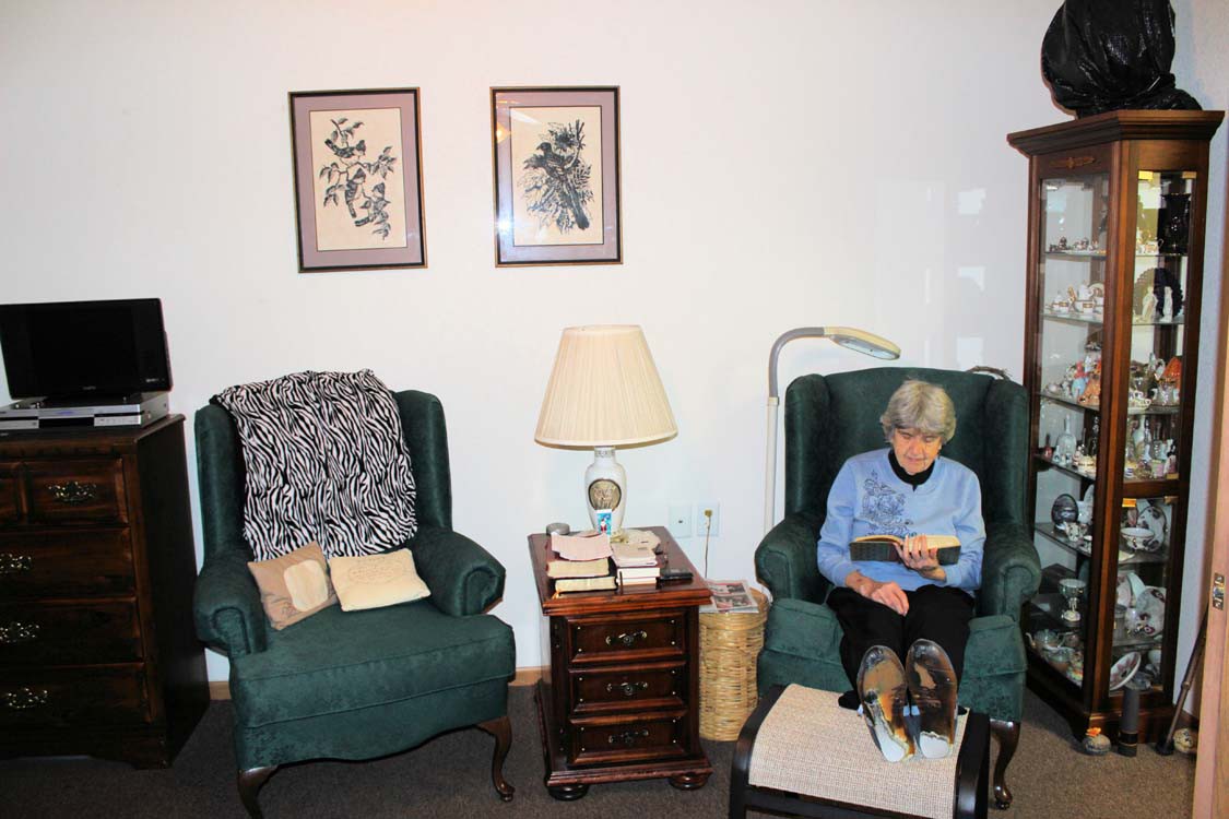 An elderly woman reading in a cozy living room with armchairs and framed art on the walls.