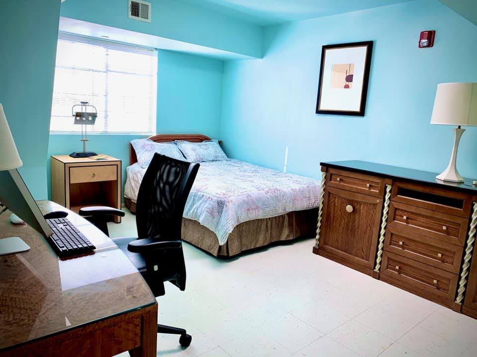 A neatly arranged bedroom with blue walls, featuring a bed, desk, chair, and dresser.