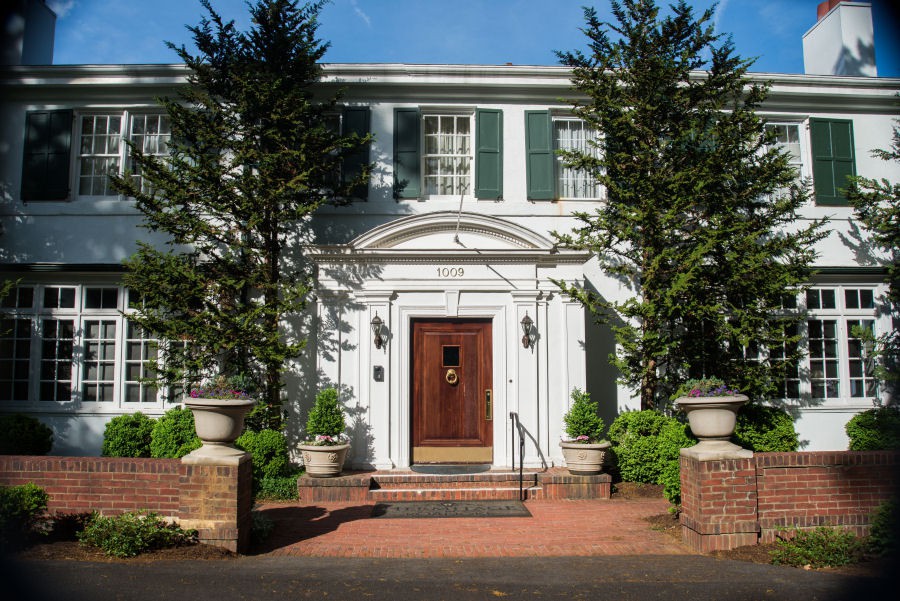 A classic two-story house with a prominent wooden door, surrounded by greenery and tall trees.