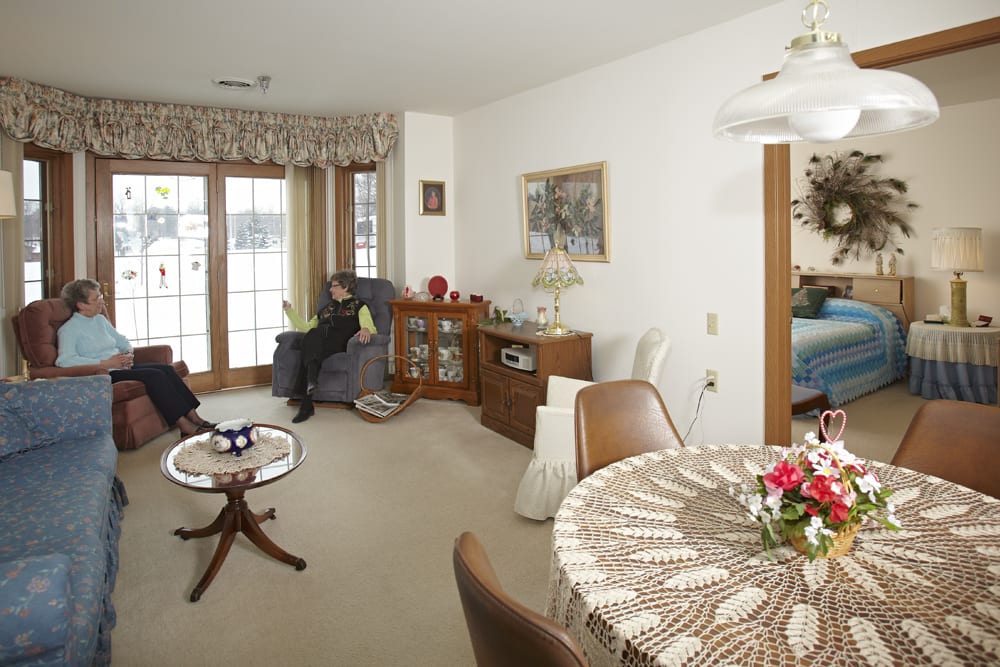 A cozy living room with a traditional interior design, featuring a dining table adorned with flowers and large windows letting in natural light.