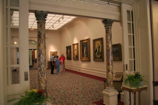 An interior view of an art gallery showcasing various paintings, with visitors admiring the artwork.