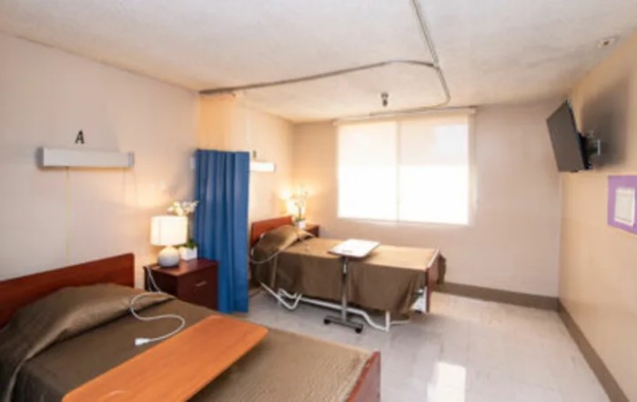 A hospital room with two beds separated by a curtain, featuring medical equipment and furniture.